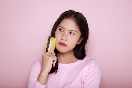 Asian woman is looking thoughtful while holding a credit card in her hand, Young woman poses alone against a light pink background, Feeling some stress about credit cards.の写真素材