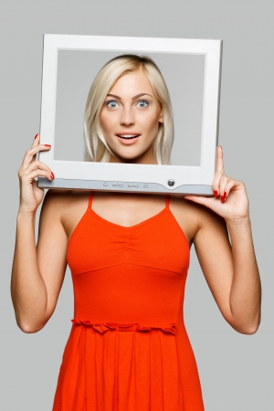 Surprised young blond female looking through the TV   computer screen frame, over gray backgroundの写真素材