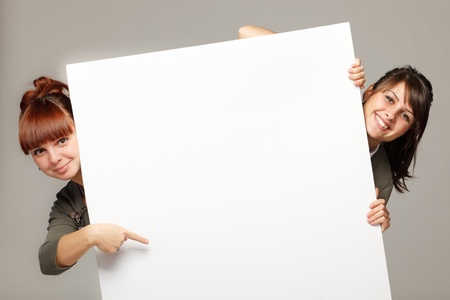 Two young women peeking over edge of blank empty paper billboard with copy space for text, over grey backgroundの写真素材