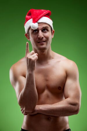 Shirtless muscular man wearing Santa hat pointing upwards, over green backgroundの写真素材