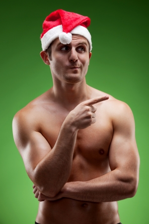 Funny image of a shirtless muscular man wearing Santa hat pointing sideways, over green backgroundの写真素材