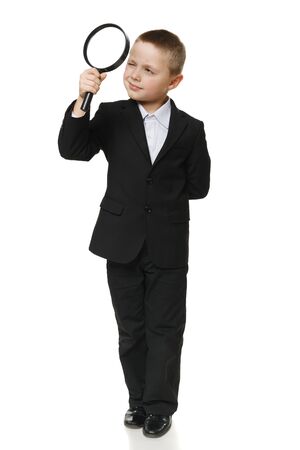 Full length of a schoolboy in black suit looking through the magnifying glass, over white backgroundの写真素材