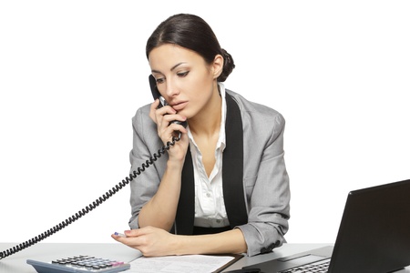 Serious business woman looking at laptop screen while talking on the phone at her work desk, isolated on white backgroundの写真素材