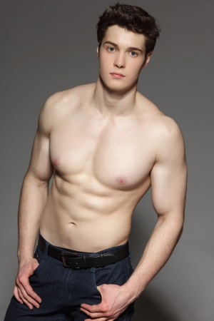 Gorgeous young man with bare torso gazing at the camera with hands in pockets, over gray backgroundの写真素材