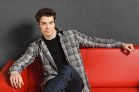 Young man relaxing sitting on the red sofa indoors with hand rested on the sofa backの写真素材
