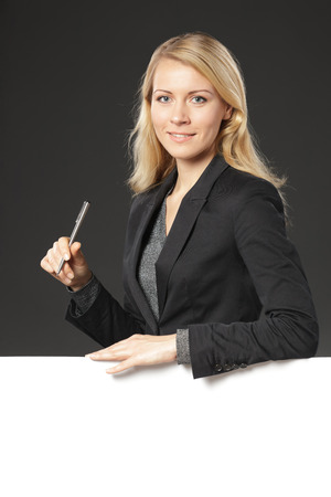 Young business woman standing behind and leaning on a white blank  holding a pen, over grey backgroundの写真素材