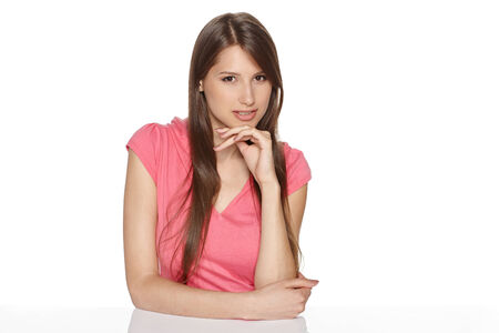 Cheerful young female sitting at white table with chin on her hand, isolated on white backgroundの写真素材