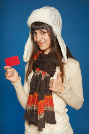 Young smiling winter woman holding credit card and showing thumb up over blue backgroundの写真素材
