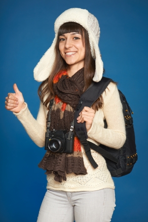 Winter girl in hat and muffler with photocamera and backpack over blue background  Happy female gesturing thumb up の写真素材