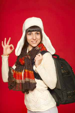 Young smiling winter woman tourist with backpack gesturing OK, over red backgroundの写真素材