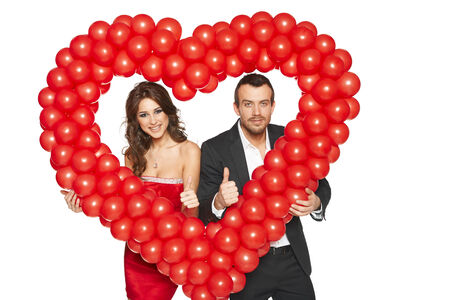 Beautiful couple looking through red balloon heart shape and gesturing thumb upの写真素材