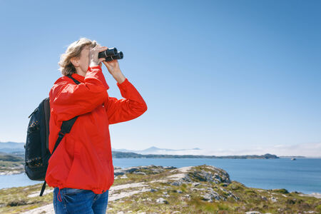 Woman tourist looking through binoculars, Norway, against blue hazy fjord backgroundの写真素材