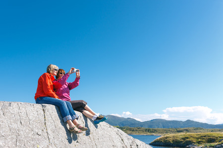 Two women sitting on stone taking photo of Norwegian landscape with fjord Kvernesfjordenの写真素材