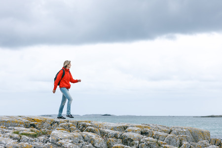 Woman hiker in red jacket and backpack walking on rock cliff against sea / ocean backgroundの写真素材