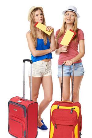 Two contemplating funky girls in shorts and straw hats standing with travel bag, holding tickets, looking away, isolated on white backgroundの写真素材