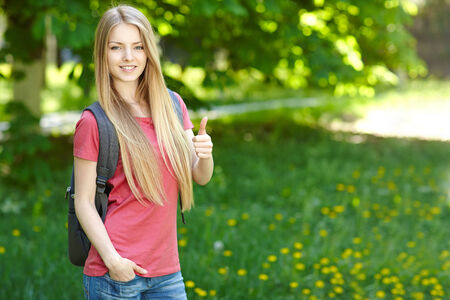 Portrait of young smiling woman outdoors with copy space. Female student with backpack on green background of city park gesturing thumb up.の写真素材