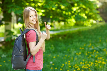 Portrait of young smiling woman outdoors with copy space. Female student with backpack on green background of city park gesturing thumb up.の写真素材
