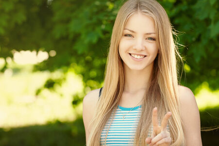 Summer girl portrait.  Closeup of a woman smiling happy on sunny summer or spring day outside in park showing victory or peace sign. Pretty young woman outdoors.の写真素材