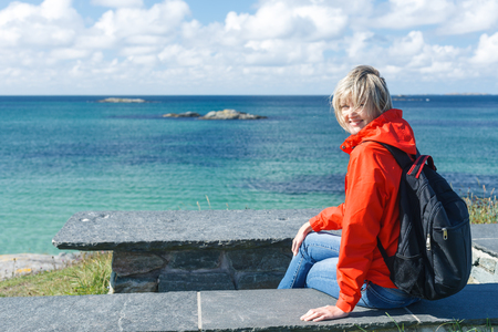 Happy woman with backpack sitting on stone bench enjoying sea / ocean / fjord view, Norwayの写真素材