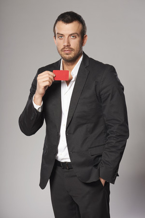 Portrait of relaxed fashionable man in suit showing red card in hand, against gray の写真素材