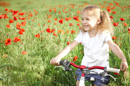 Happy smiling little girl standing with bicycle on the poppy meadow laughing looking awayの写真素材