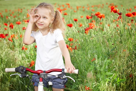 Happy smiling little girl standing with bicycle on the poppy meadow looking away through circled fingersの写真素材