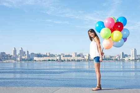 Summer holiday, celebration and present concept: happy young woman holding colorful balloons behind back, standing in full length, urban scene,city view, outdoorsの写真素材