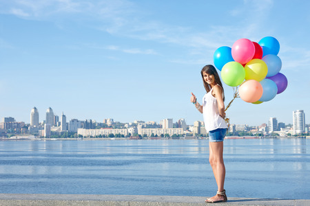 Summer holiday, celebration and present concept: happy young woman holding colorful balloons behind back and gesturing thumb up, standing in full length, city view, outdoorsの写真素材