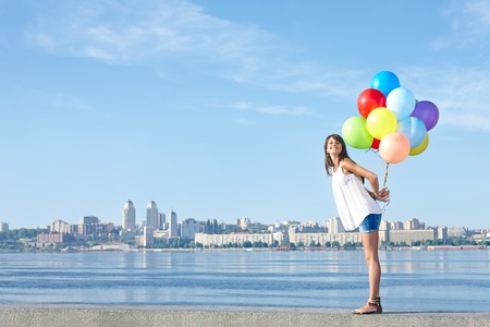Summer holiday, celebration and present concept: happy young woman holding colorful balloons behind back, standing in full length, city view, outdoorsの写真素材