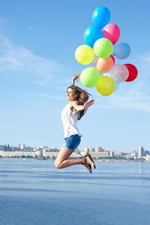 Excited young woman with colorful balloons jumping over city backgroundの写真素材