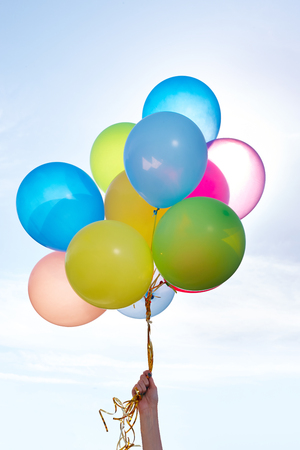 Hand holding bunch of colorful balloons in blue sky, backlit by sunの写真素材