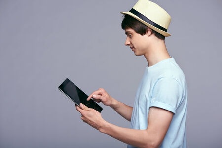 Tablet computer. Side view of man in blue tshirt and straw hat using digital tablet isolated on gray background.の写真素材