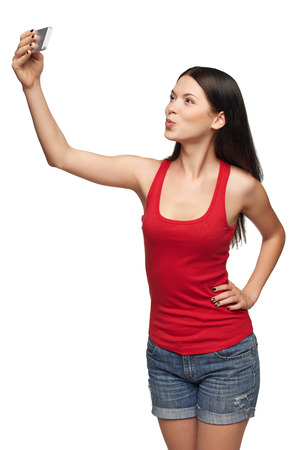 Happy young girl making kiss while taking pictures of herself through cellphone, over white backgroundの写真素材