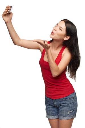 Happy young girl making kiss while taking pictures of herself through cellphone, over white backgroundの写真素材