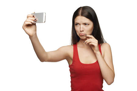 Young girl pulling discontent face while taking pictures of herself through cellphone, over white backgroundの写真素材
