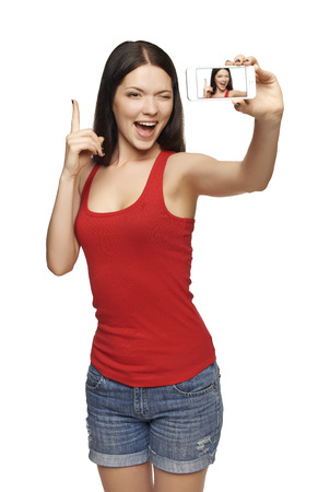 Happy excited young woman with finger up, while taking pictures of herself through cell phone, over white backgroundの写真素材