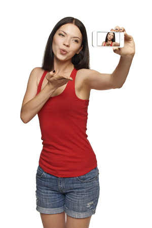 Happy young girl making kiss while taking pictures of herself through cellphone, over white backgroundの写真素材