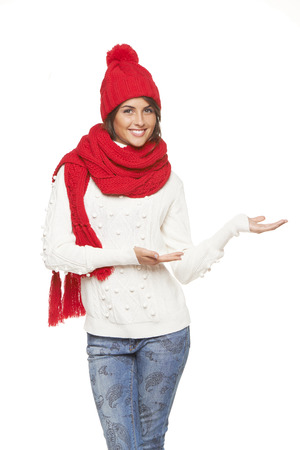 Winter, christmas, holidays concept. Smiling beautiful woman in warm winter red hat and scarf showing open hand palm with copy space for product or text, over gray backgroundの写真素材