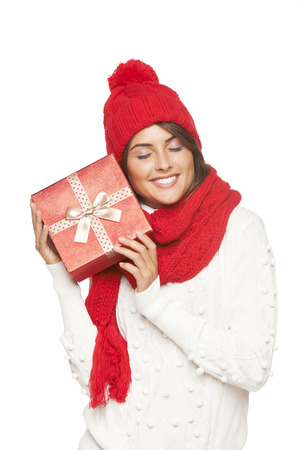 Christmas gift. Smiling beautiful woman in winter red hat and scarf envisioning with closed eyes what is in the gift box, over white backgroundの写真素材