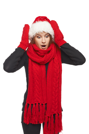 Surprised shocked Christmas woman wearing santa hat and red scarf and mittens with hands on head, looking down, over white backgroundの写真素材