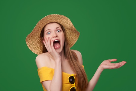 Shocked surprised woman wearing bright yellow dress and summer straw hat showing open hand palm with copy space, over green backgroundの写真素材