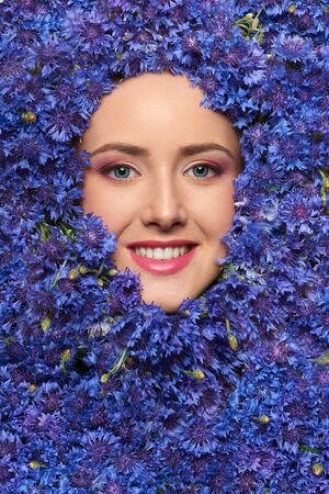 Closeup of happy smiling beauty woman face with perfect skin and professional makeup in blue bachelor's-button cornflower flowers.の写真素材