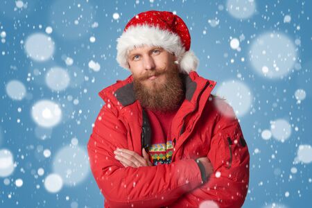 Bearded man in red winter jacket and Santa hat, over snow backgroundの写真素材