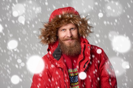 Closeup portrait of bearded man in red winter jacket with hood on, over snow backgroundの写真素材