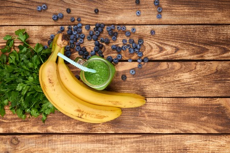 Top view of jar tumbler mug with green smoothie drink and a bundle of fresh parsley, banana and blueberries on wooden tableの写真素材