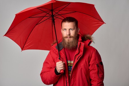 Portrait of bearded man in red winter jacket standing with umbrella, over grey background