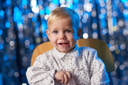 Smiling toddler child sitting on the wooden chair over blue holidays bokeh backgroundの写真素材