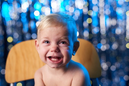 Crying toddler child sitting on the wooden chair over blue holidays bokeh background