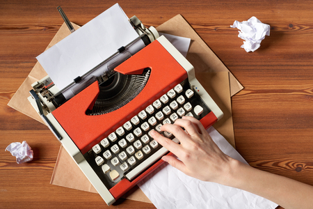 Top view of woman hand typing on red vintage typewriter with white blank paper sheet on wooden tableの写真素材