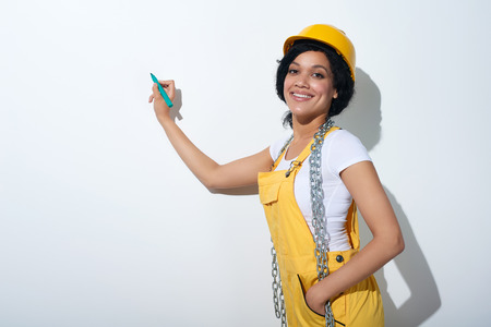 Happy smiling mixed race construction woman wearing yellow protect helmet and overall holding a pen going to write on white blank copy spaceの写真素材